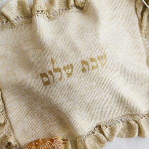 Peut inclure: Une housse à challah tricotée de couleur crème avec une bordure à volants et le texte hébreu "Shabbat Shalom" brodé en fil doré scintillant. Le tissu présente un léger éclat métallique sur tout le tricot, créant un aspect texturé.