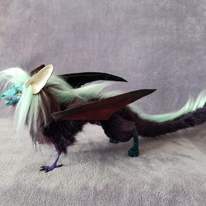 May include: A handmade fantasy creature plush toy. The plush has a purple body, teal and white fur, and burgundy wings. The creature has a teal face, silver ears, and a long tail. The plush is posed on a grey surface.