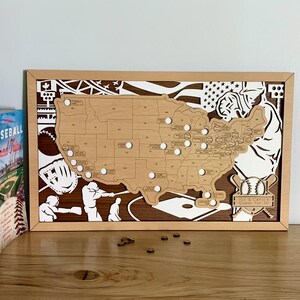 May include: A rectangular wooden wall art piece featuring a laser-cut map of the United States with circular cutouts for baseball stadiums. The design includes white silhouettes of baseball players and equipment, with a plaque reading Vic & Carol's Quest for 30.