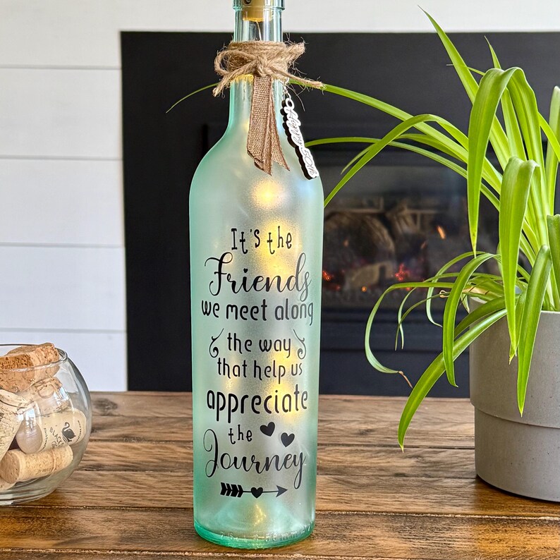 May include: A light green glass bottle with a cork stopper and string decorations. The bottle features the text "It's the Friends we meet along the way that help us appreciate the Journey". A small light illuminates the inside.