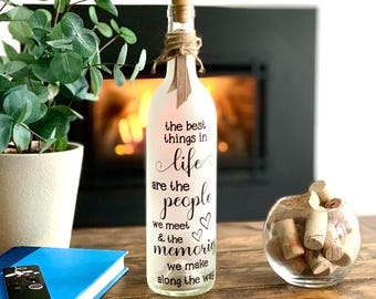 Könnte beinhalten: Eine dekorative Glasflasche mit Korken, Bindfaden und einem Band. Auf der Flasche steht der Text "the best things in life are the people we meet & the memories we make along the way". Ein blaues Buch und eine Fernbedienung sind im Vordergrund.