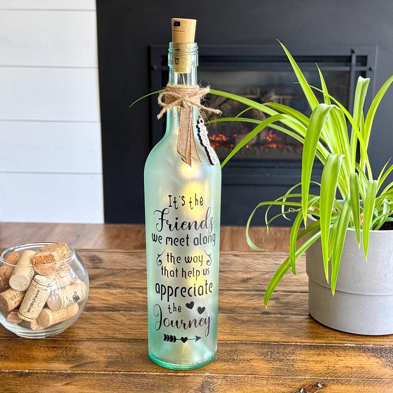 May include: A light-green glass bottle with a cork stopper and string decorations. The bottle has the text "It's the Friends we meet along the way that help us appreciate the Journey." The bottle is illuminated from within.