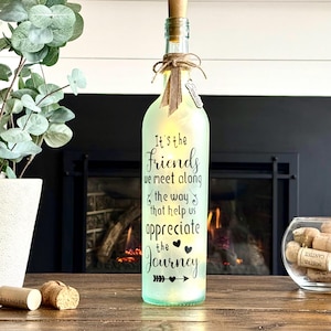 May include: A decorative, illuminated wine bottle with a cork stopper. The bottle is frosted green and features the text "It's the Friends we meet along the way that help us appreciate the Journey." A small charm hangs from a twine bow.