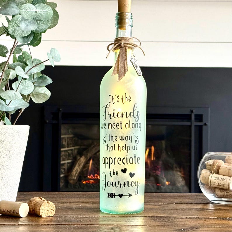 May include: A decorative, illuminated wine bottle with a cork stopper. The bottle is frosted green and features the text "It's the Friends we meet along the way that help us appreciate the Journey." A small charm hangs from a twine bow.