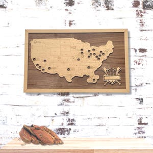 May include: A rectangular wooden wall display featuring a light-toned map of the United States mounted on a dark wood background. The map includes circular cutouts for baseballs and a decorative emblem with two crossed bats and a baseball.
