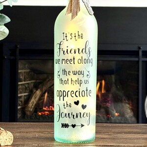 May include: A decorative, illuminated glass bottle with the text "It's the Friends we meet along the way that help us appreciate the Journey." The bottle has a light green tint and is decorated with a bow.