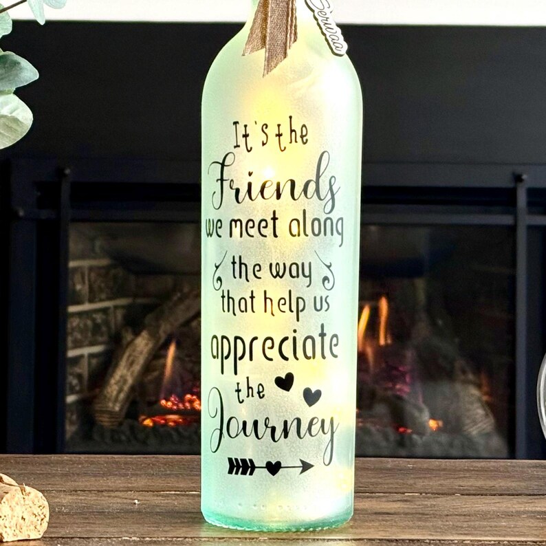 May include: A decorative, illuminated glass bottle with the text "It's the Friends we meet along the way that help us appreciate the Journey." The bottle has a light green tint and is decorated with a bow.