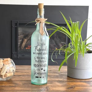 May include: A decorative glass bottle with a cork stopper, decorated with a twine bow and a feather. The bottle has the text "It's the Friends we meet along the way that help us appreciate the Journey".
