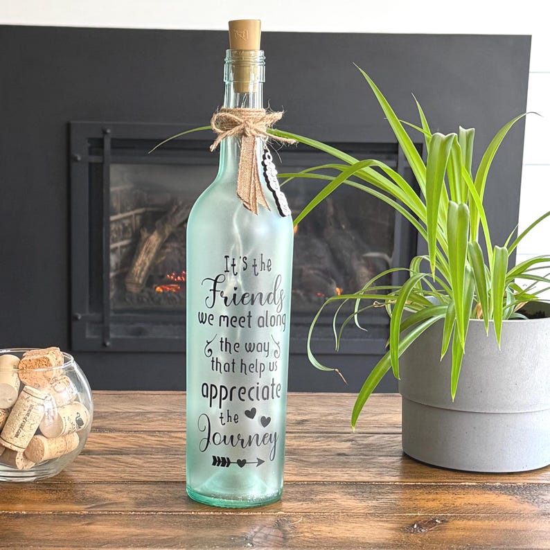 May include: A decorative glass bottle with a cork stopper, decorated with a twine bow and a feather. The bottle has the text "It's the Friends we meet along the way that help us appreciate the Journey".