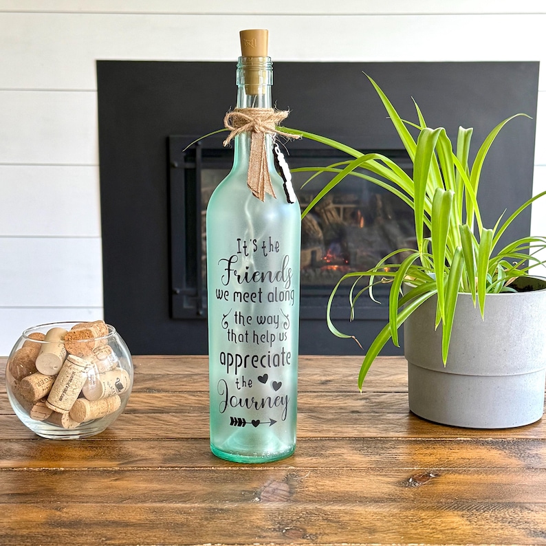 May include: A light green glass bottle with a cork stopper and a jute twine bow. The bottle has the text "It's the Friends we meet along the way that help us appreciate the Journey". A small glass bowl with corks is to the left.