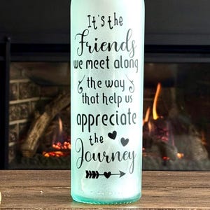 May include: A light green glass bottle with the text "It's the Friends we meet along the way that help us appreciate the Journey" in black script. The bottle is on a wooden surface with a fireplace in the background.