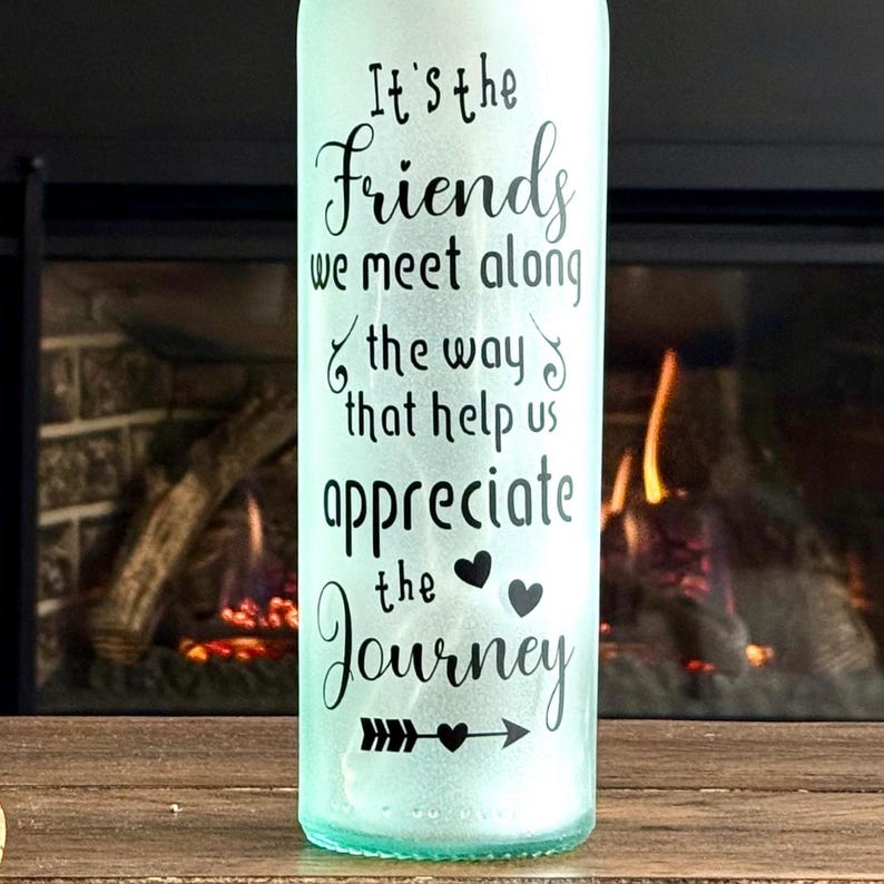 May include: A light green glass bottle with the text "It's the Friends we meet along the way that help us appreciate the Journey" in black script. The bottle is on a wooden surface with a fireplace in the background.