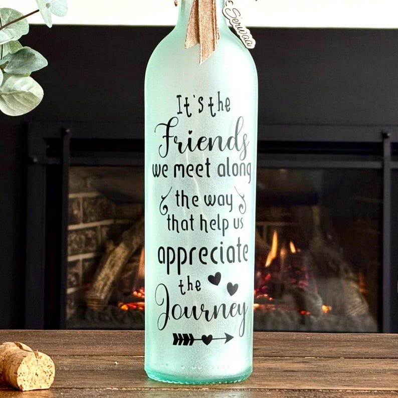 May include: A frosted glass bottle with the text "It's the Friends we meet along the way that help us appreciate the Journey." The bottle has a burlap ribbon and a cork. The background includes a fireplace.