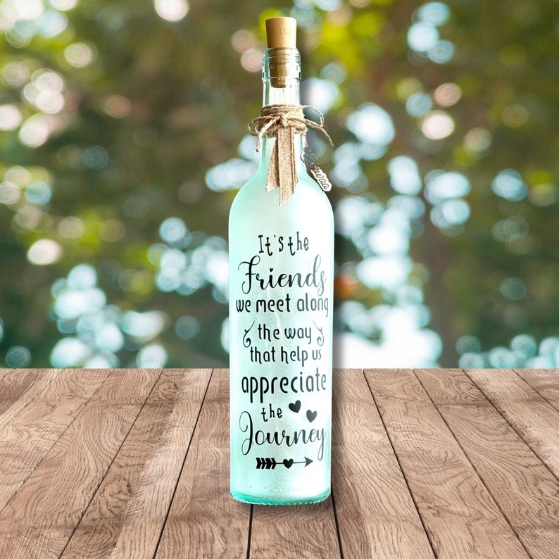 May include: A frosted glass bottle with a cork stopper, tied with twine and a small tag. The bottle is light blue and has the text "It's the Friends we meet along the way that help us appreciate the Journey".
