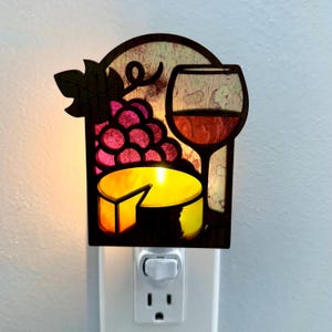 May include: A decorative night light featuring a wine theme. The design includes a wine glass, a bunch of grapes, and a wheel of cheese. The light is plugged into a wall outlet and has a switch to turn it on and off.