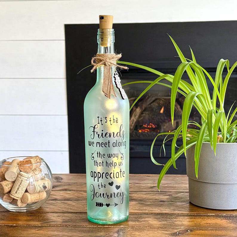 May include: A decorative, light-up glass bottle with a cork stopper. The bottle is frosted green and features the text "It's the Friends we meet along the way that help us appreciate the Journey." A small butterfly and twine decorate the neck.