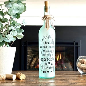 May include: A decorative glass bottle with a cork and twine bow. The bottle is frosted and has the text "It's the Friends we meet along the way that help us appreciate the Journey" in black script. A small metal tag reads "Serwaa".
