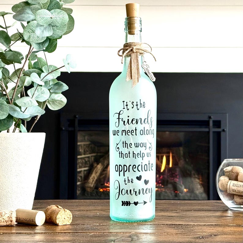 May include: A decorative glass bottle with a cork and twine bow. The bottle is frosted and has the text "It's the Friends we meet along the way that help us appreciate the Journey" in black script. A small metal tag reads "Serwaa".