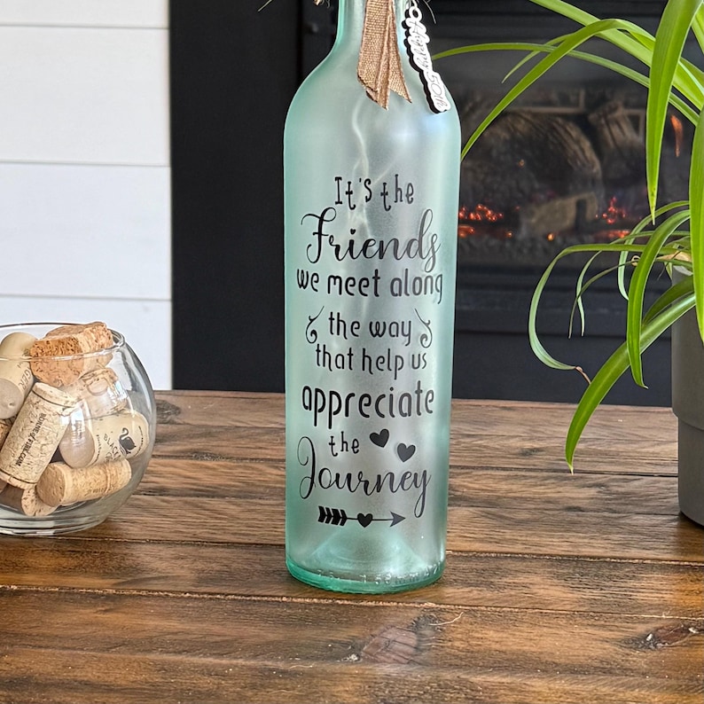 May include: A light green glass bottle with the text "It's the Friends we meet along the way that help us appreciate the Journey." The bottle has a burlap ribbon and a small charm attached to the top. A glass bowl of corks is in the background.