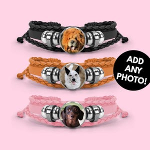 May include: Three braided bracelets with dog portraits. The top bracelet is black, the middle is tan, and the bottom is pink. Each bracelet has a silver clasp and a round photo of a dog. The text "ADD ANY PHOTO!" is on a black circle.