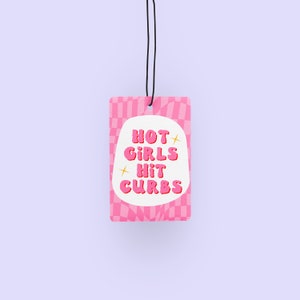 May include: Pink car air freshener with a white oval containing the text "HOT GIRLS HIT CURBS" in pink bubble letters. The background is a pink and white checkered pattern.