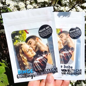 Peut inclure: Deux d&eacute;sodorisants de voiture fantaisie dans un emballage blanc. Chacun pr&eacute;sente une photo d'un couple s'embrassant. Le texte sur l'emballage comprend "Binge Greetings" et "Novelty Car Air Freshener". Un autocollant noir dit "REUSE ME!" avec des instructions.