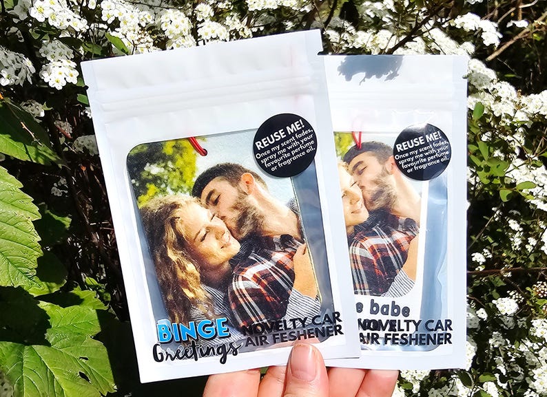 Peut inclure: Deux d&eacute;sodorisants de voiture fantaisie dans un emballage blanc. Chacun pr&eacute;sente une photo d'un couple s'embrassant. Le texte sur l'emballage comprend "Binge Greetings" et "Novelty Car Air Freshener". Un autocollant noir dit "REUSE ME!" avec des instructions.
