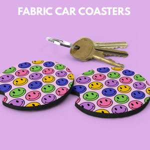 May include: Two fabric car coasters with a pink background and a pattern of colorful smiley faces. The coasters are next to a set of keys. The text "FABRIC CAR COASTERS" is at the top.