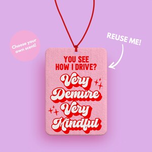 Peut inclure: Diffuseur de parfum de voiture rose avec le texte "YOU SEE HOW I DRIVE? Very Demure Very Mindful" en rouge et blanc dans une police r&eacute;tro. Le diffuseur est suspendu &agrave; une ficelle rouge.