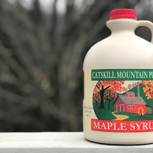 May include: A white plastic jug with a red lid and handle. The jug contains Catskill Mountain Pure Maple Syrup. The label features a red barn with a green roof and a yellow path leading to it. The barn is surrounded by trees with red and orange leaves. The label also includes the text "Catskill Mountain Pure Maple Syrup".