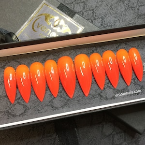 Puede incluir: Un juego de diez uñas postizas largas y puntiagudas, con degradado naranja y rojo. Las uñas están dispuestas en una fila dentro de una caja negra. El nombre de la marca "venomnails.com" está impreso en la caja. Un logotipo dorado es visible en la tapa.