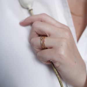 May include: Three gold rings with small pink gemstones. The rings are stacked on a finger.