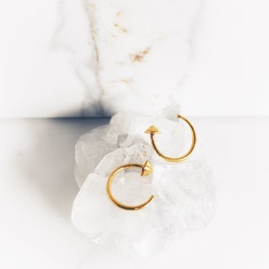 May include: Two gold-coloured hoop earrings with a geometric design. Each earring has a small, triangular accent at the top. The earrings are displayed on a clear, crystalline surface against a white background.