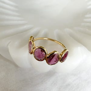 Op de afbeelding: Een gouden ring met vijf ovale, roze edelstenen. De stenen zijn in een rij op de ringband geplaatst. De ring wordt gepresenteerd op een wit, gestructureerd oppervlak, waardoor het ontwerp en de kleur van de ring worden benadrukt.