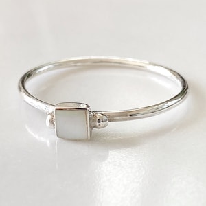 Può includere: Un anello d'argento con una pietra quadrata bianca incastonata al centro. L'anello ha due piccole perle d'argento su entrambi i lati della pietra.