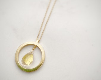 May include: A gold-colored necklace featuring a circular pendant with a textured finish. A teardrop-shaped, yellow gemstone hangs within the circle. The necklace chain is delicate and gold-toned, set against a white background.