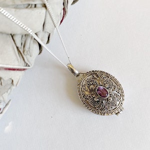 Puede incluir: Un collar de plata con un colgante de filigrana con una piedra preciosa púrpura. El colgante tiene forma ovalada y un diseño delicado e intrincado.