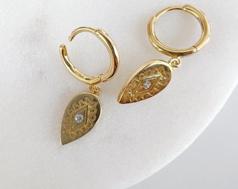 May include: A pair of gold-colored hoop earrings with teardrop-shaped pendants. Each pendant features a detailed geometric design with a small, clear gemstone in the center. The earrings are displayed on a white surface.