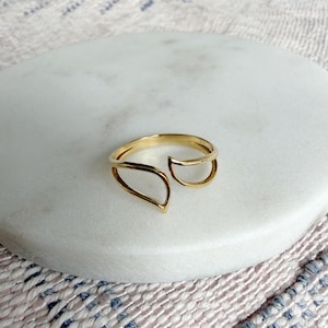 May include: A gold ring with an unusual open design. The ring has a curved band with two open, leaf-like shapes. The ring is set on a white marble surface, with a patterned fabric in the background.