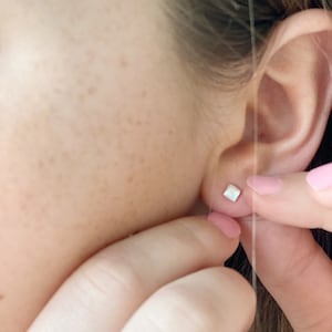 Könnte beinhalten: Nahaufnahme eines Ohrs mit einem kleinen, quadratischen Ohrring. Der Ohrring ist weiß und hat einen dezenten Schimmer.