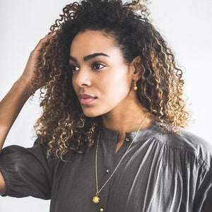 May include: A woman with curly brown hair wearing a gray shirt and gold jewelry. She is wearing a gold necklace with a flower pendant and gold earrings with flower accents.