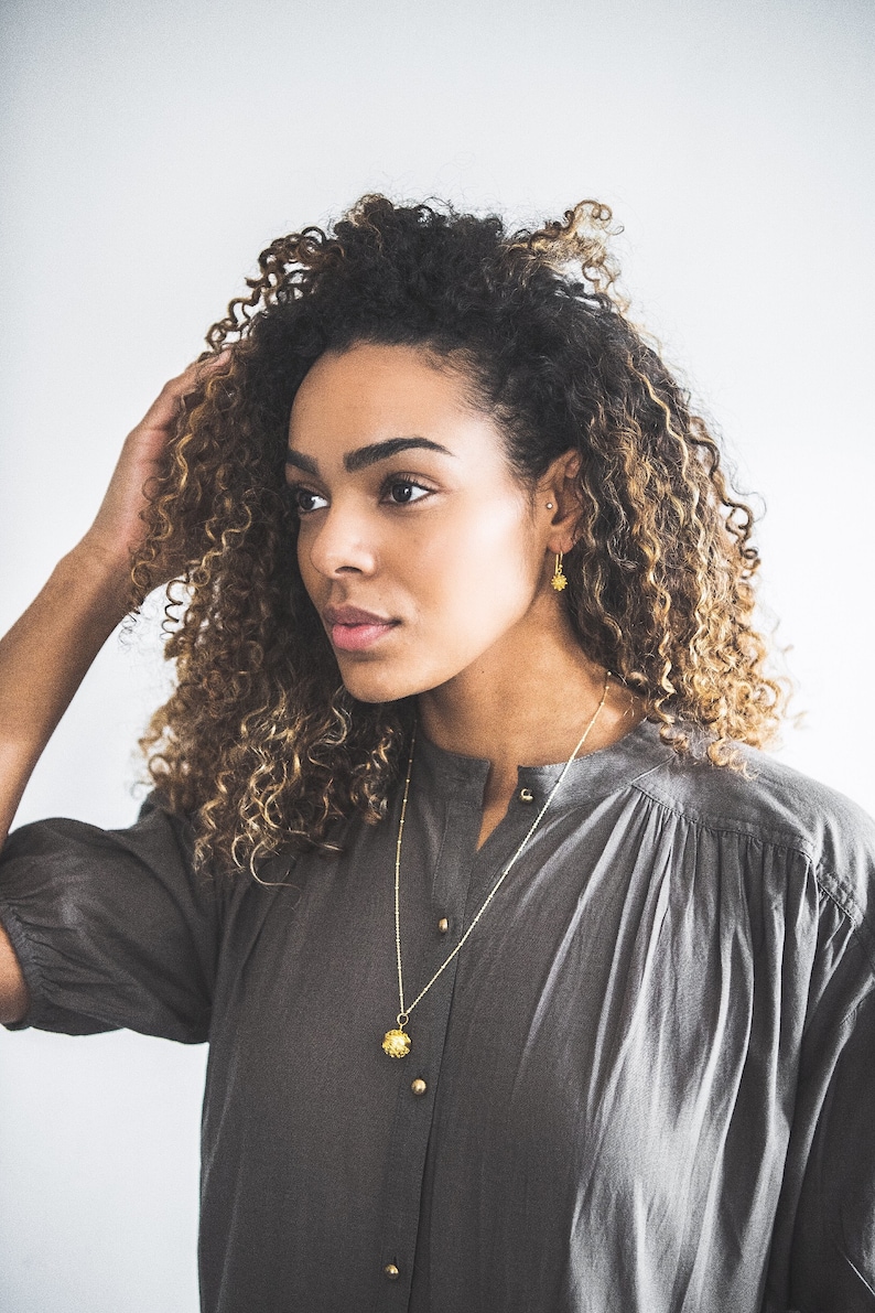 May include: A woman with curly brown hair wearing a gray shirt and gold jewelry. She is wearing a gold necklace with a flower pendant and gold earrings with flower accents.