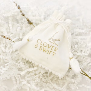 May include: White drawstring bag with a gold logo that says "Clover & Swift" and a small gold bird design. The bag is sitting on white crinkle paper.