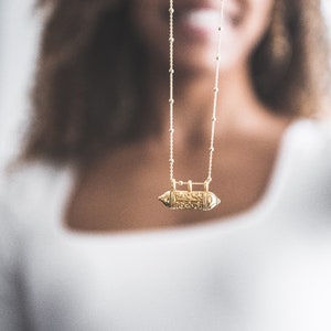May include: A gold chain necklace with a small, ornate, gold pendant in the shape of a capsule.
