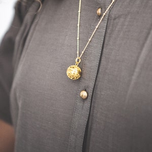 May include: A gold chain necklace with a round, ornate pendant. The necklace is worn by a person wearing a gray shirt.