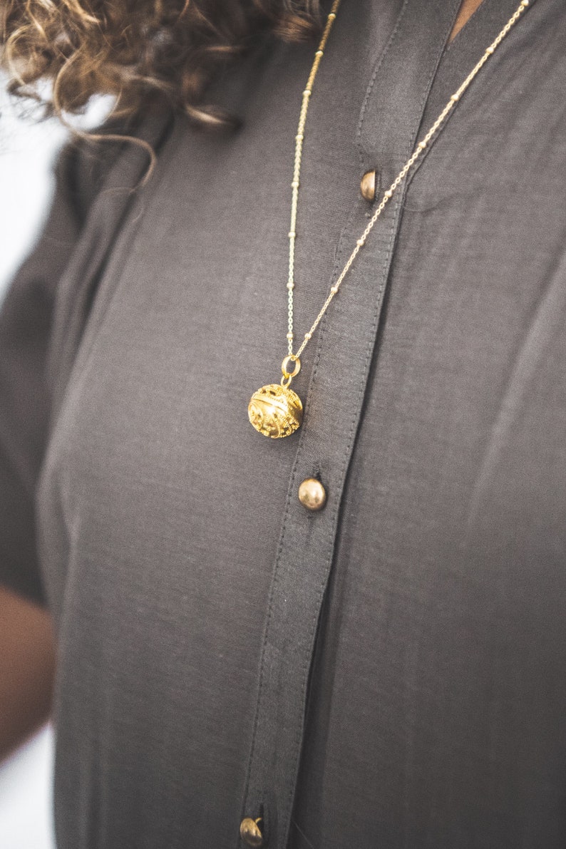 May include: A gold chain necklace with a round, ornate pendant. The necklace is worn by a person wearing a gray shirt.