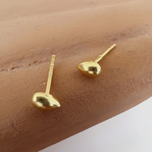 May include: A pair of gold-coloured stud earrings. Each earring has a small, rounded, teardrop-shaped design. The earrings are set against a brown surface, highlighting their metallic sheen. These earrings are a simple yet elegant jewellery item.
