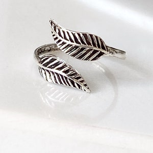 Op de afbeelding: Een zilverkleurige ring met een bladvormgeving. De ring heeft twee gedetailleerde bladvormen die zich om de vinger wikkelen. De bladeren hebben getextureerde nerven en een licht geoxideerde afwerking, wat het een vintage uitstraling geeft. De ring staat tegen een witte achtergrond.
