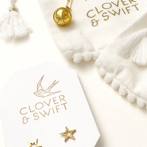 May include: A white pouch with gold pom-poms and the text "Clover & Swift" with a gold geometric bird design. A gold necklace with a gold ball pendant is in the pouch. A white card with the text "Clover & Swift" and a gold geometric bird design is on a white surface. Two gold star-shaped earrings are on the card.