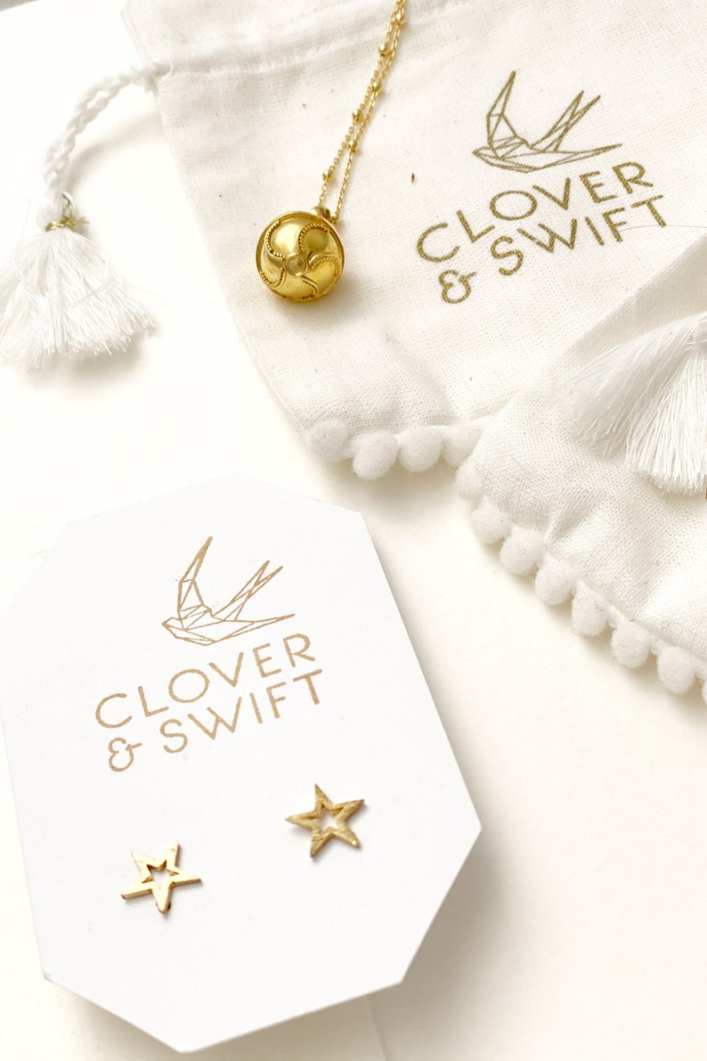 May include: A white pouch with gold pom-poms and the text "Clover & Swift" with a gold geometric bird design. A gold necklace with a gold ball pendant is in the pouch. A white card with the text "Clover & Swift" and a gold geometric bird design is on a white surface. Two gold star-shaped earrings are on the card.